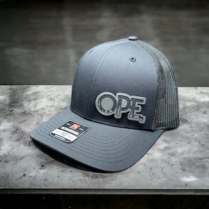 May include: A gray and black trucker hat with a gray embroidered logo that says "OPE" on the front. The hat has a mesh back and a curved brim.