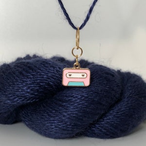 Puede incluir: Un charm de cinta de casete rosa y azul con detalles dorados y dos pequeños corazones como ojos. El charm está unido a un anillo de salto dorado y un cordón negro.