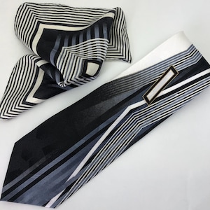 May include: A black and white patterned tie with a black and white patterned pocket square. The tie has a geometric design with a silver label.