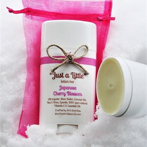 May include: A white lotion bar with a pink and white label that reads "Just a little lotion bar Japanese Cherry Blossom". The label also includes the ingredients and contact information for the maker. The lotion bar is sitting on a white surface with a pink mesh bag behind it.
