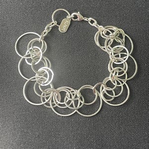 May include: Silver-toned bracelet featuring a series of interconnected circles. The bracelet has a lobster clasp closure and a small tag with the brand name. The design is modern and geometric, suitable for everyday wear.