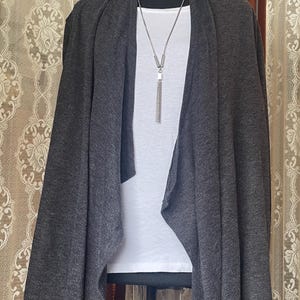 May include: A dark grey draped cardigan over a white t-shirt, displayed on a mannequin. A silver necklace with a tassel hangs from the neckline. The cardigan has a relaxed fit and a waterfall front. The background features a patterned curtain.