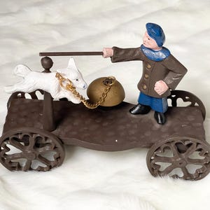 Può includere: Statuetta decorativa raffigurante un carretto marrone con ruote a raggi, un cane bianco e una persona con cappello e cappotto blu. Il cane è attaccato al carretto con una catena d'oro. La persona tiene un bastone.