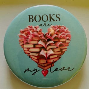 May include: Round, light blue button with the words "BOOKS are my love." A heart shape made of stacked books with pink pages and red covers is in the center. A small red heart is below the word "my".