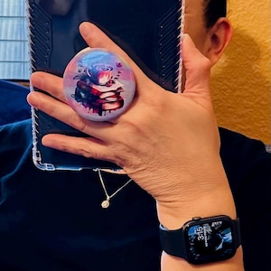 May include: A black e-reader with a clear case and a colorful phone grip with a floral design. The e-reader is being held by a person wearing a black shirt and a black Apple Watch with a digital clock face that reads 3:15.