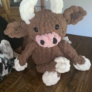 Könnte beinhalten: Ein braunes und weißes Plüsch-Kuh-Spielzeug mit einer rosa Nase und schwarzen Augen. Die Kuh sitzt auf einer Holzoberfläche.