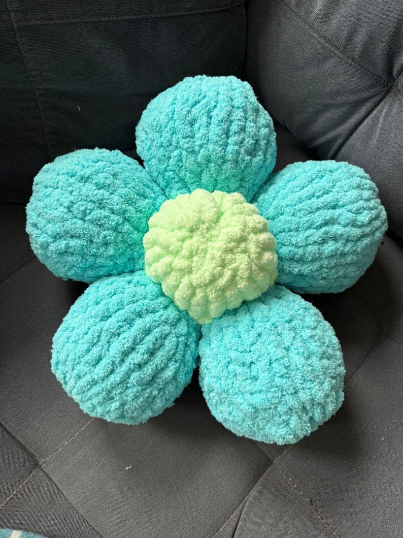 May include: A light blue and green crocheted flower-shaped pillow. The pillow is made of soft, fluffy yarn and has a five-petal design.