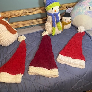 May include: Five knitted items, including three red and white Santa hats, a white snowman with an orange scarf, and a green and blue snowman with a blue scarf. The items are arranged on a blue blanket.