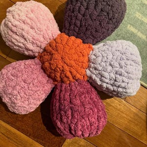 May include: A large, crocheted flower-shaped pillow with five petals in shades of pink, purple, and orange. The pillow is made of a soft, fluffy yarn and has a textured surface.