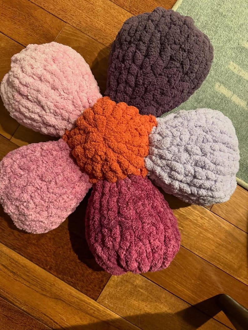 May include: A large, crocheted flower-shaped pillow with five petals in shades of pink, purple, and orange. The pillow is made of a soft, fluffy yarn and has a textured surface.