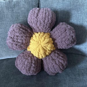 May include: A plush flower-shaped pillow with a purple outer layer and a yellow center. The pillow is sitting on a gray couch.