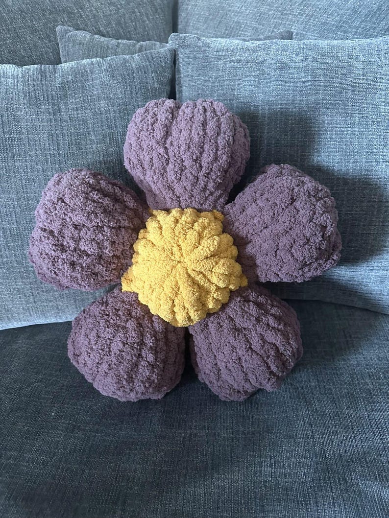 May include: A plush flower-shaped pillow with a purple outer layer and a yellow center. The pillow is sitting on a gray couch.