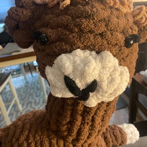 May include: A brown crocheted llama with white fur around the nose and black eyes. The llama has a brown fluffy mane.