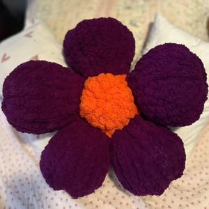 May include: A large purple flower-shaped pillow with an orange center. The pillow is made of a soft, fuzzy material.