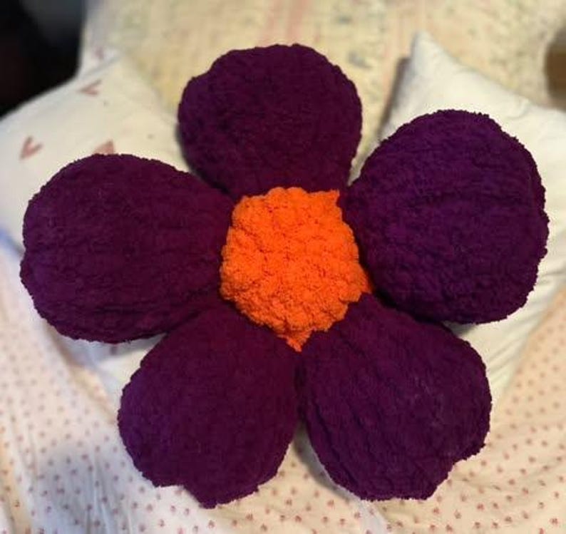 May include: A large purple flower-shaped pillow with an orange center. The pillow is made of a soft, fuzzy material.