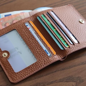 Op de afbeelding: Een bruine leren portemonnee, opengeklapt om creditcards, een identiteitskaart en gevouwen bankbiljetten te laten zien. De portemonnee heeft een drukknoopsluiting en een doorzichtig venster voor een ID. Op een houten oppervlak.