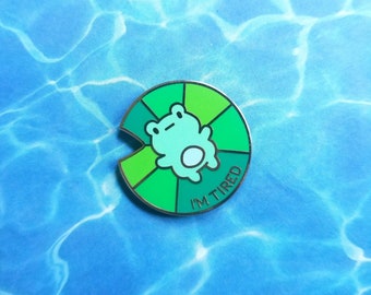 May include: A round enamel pin featuring a light green frog on a green lily pad design. The pin has the text "I'M TIRED" in white. The pin is set against a blue background.