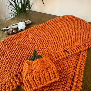 Könnte beinhalten: Eine grob gestrickte orangefarbene Decke mit einer passenden orangefarbenen Mütze mit grünem Stiel. Die Decke hat eine sichtbare Rippenstruktur und ein kleines braunes rechteckiges Etikett an der Seite. Das Set besteht aus dickem Garn.