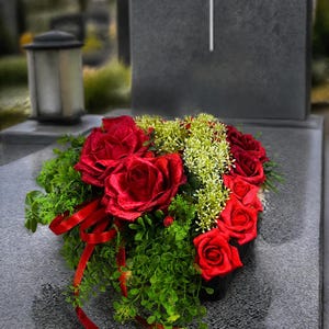 Könnte beinhalten: Ein Blumenarrangement mit dunkelroten Rosen und kleinen weißen Füllblumen inmitten von grünem Blattwerk. Der Strauß ist mit dünnen roten Bändern verziert und liegt auf einer dunkelgrauen Steinfläche vor einem Grabstein mit einem weißen Kreuz.