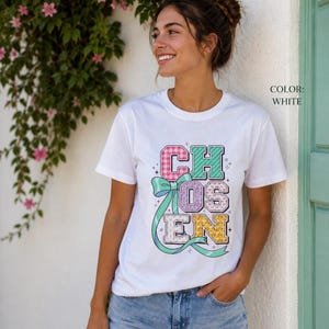 Può includere: Una t-shirt bianca a maniche corte con la parola CHOSEN in lettere maiuscole colorate e a motivi. Le lettere sono decorate con un fiocco a nastro azzurro e piccole stelle. La t-shirt ha un girocollo e una vestibilità standard.
