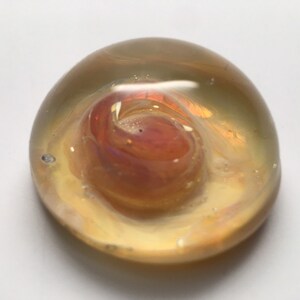 Puede incluir: Un pisapapeles redondo de vidrio transparente con un diseño de remolino opaco, rosa y naranja en su interior.