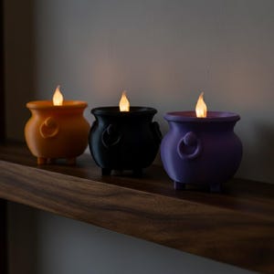Könnte beinhalten: Drei LED-Kerzen in Kesselform in Orange, Schwarz und Lila, jede mit einer flackernden Flamme. Die Kerzen haben ein Griffdetail und werden auf einem Holzregal ausgestellt. Perfekt für Halloween-Dekorationen.