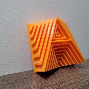 Pu&ograve; includere: Una lettera A arancione stampata in 3D, realizzata con triangoli sovrapposti, &egrave; appoggiata a un muro bianco.
