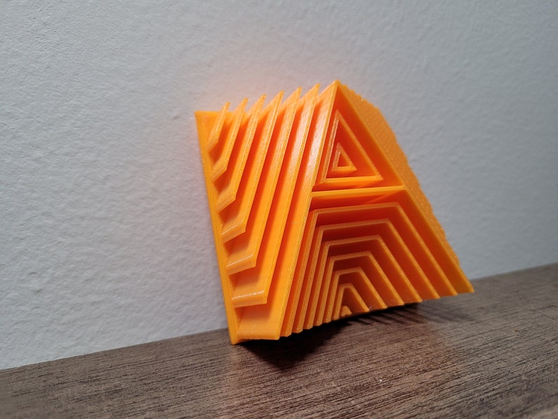 Pu&ograve; includere: Una lettera A arancione stampata in 3D, realizzata con triangoli sovrapposti, &egrave; appoggiata a un muro bianco.