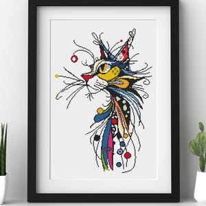 May include: A framed cross-stitch pattern featuring a stylised cat head in profile. The cat is rendered with vibrant yellow, blue, red, and black thread, incorporating intricate line work, geometric shapes, and small colourful dots throughout the design.