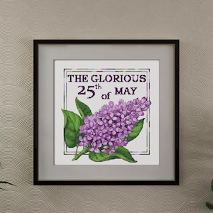 Puede incluir: Un bordado enmarcado que representa una flor de lilas con hojas verdes. El texto "THE GLORIOUS 25th of MAY" se muestra encima de la flor. La obra de arte está enmarcada sobre una pared texturizada neutra.