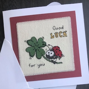 May include: A white greeting card with a square cross-stitched insert featuring a red ladybird on a green four-leaf clover. The text reads Good LUCK for you in black and yellow thread. The card is accompanied by a matching white envelope.