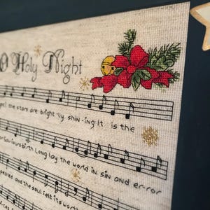 May include: A cross-stitch pattern of the song "O Holy Night" with musical notes and a red poinsettia with a gold bell.