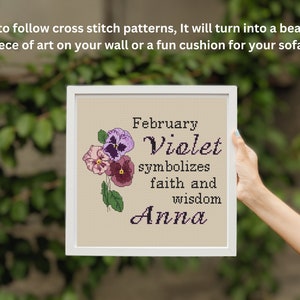 K&ouml;nnte beinhalten: Ein gerahmtes Kreuzstichmuster mit einem floralen Design und dem Text "Februar Veilchen symbolisiert Glauben und Weisheit Anna".