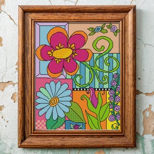May include: A framed, colorful floral cross-stitch artwork. The design features large flowers in pink, orange, and blue, with green vines and leaves. The background is divided into geometric sections with various patterns and colors. The frame is made of wood.