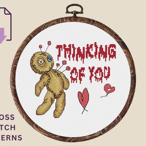 以下が含まれることがあります： クロスステッチパターン。ピンの付いたブードゥー人形、「THINKING OF YOU」の文字、2つの赤いハートが描かれています。木製の刺繍枠にデザインされており、下部には「CROSS STITCH PATTERNS」の文字があります。