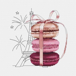 Op de afbeelding: Drie roze en bruine macarons die met een roze lint en strik aan elkaar zijn gebonden. De macarons zijn op elkaar gestapeld. Op de achtergrond is een zwart-witte omtrek van de Eiffeltoren te zien.