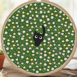 Puede incluir: Un bastidor de bordado de punto de cruz con un gato negro asomando sobre un fondo verde con flores blancas y amarillas. El aro de madera enmarca el diseño bordado, creando una pieza artesanal y encantadora.