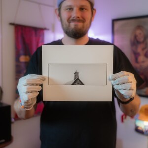 May include: A person is holding a white mounted print of a black and white photograph of a building with a spire. The building is against a light grey sky.