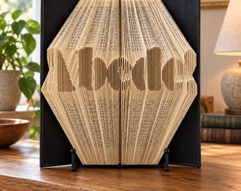 May include: A folded book art sculpture featuring the letters 'abcde' created by precisely folding the individual pages of a book. The sculpture is displayed upright on a black metal stand, set against a black backing board, showcasing the intricate paper edges.