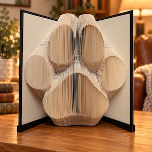 Peut inclure: Une sculpture de livre avec des pages pliées formant la silhouette d'une empreinte de patte. Le livre est ouvert avec des couvertures noires de chaque côté, mettant en valeur la technique de pliage de papier utilisée pour créer ce motif.