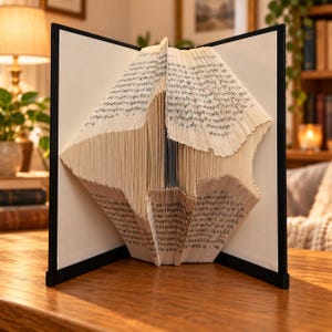 May include: A folded book sculpture featuring pages intricately creased to form a geometric diamond shape. The open book is held upright by a black frame, displaying the printed text on the paper edges, creating a structured, three-dimensional art piece.