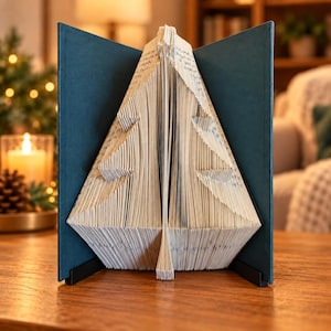 Könnte beinhalten: Eine Buchskulptur mit gefalteten Seiten, die die Form eines Weihnachtsbaums bilden. Das Buch hat dunkelblau-grüne Einbände, die offen stehen und die weißen, gefalteten Papierseiten einrahmen. Die Struktur steht auf einem schwarzen Metallständer.