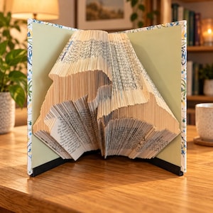 May include: A book sculpture featuring folded pages that form the silhouette of a dolphin. The book is open, displaying the intricate paper folding technique with the text of the pages visible along the edges of the sculpted dolphin shape.