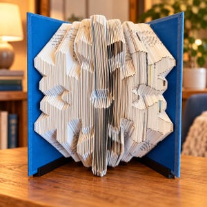 Könnte beinhalten: Eine Buchskulptur mit gefalteten Seiten, die ein symmetrisches Schneeflockenmuster bilden. Das Buch wird durch leuchtend blaue Einbände auf beiden Seiten offen gehalten, was eine starre Struktur erzeugt. Die weißen Papierseiten sind präzise gefaltet.