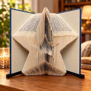 May include: A book sculpture featuring pages folded to create a prominent five-pointed star shape. The book has a blue cover and is held open by two black metal stands, displaying the intricate, layered paper edges of the star design clearly.