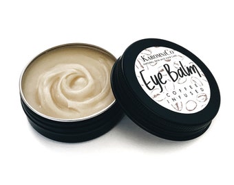 May include: A small black tin with a white label that reads "Karmic Co. Eye Balm Coffee Infused". The tin is open and the lid is next to it. The tin contains a light brown balm.