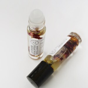 Pode incluir: Dois frascos roll-on de &oacute;leo essencial de vidro transparente com tampas pretas e r&oacute;tulos brancos. Um frasco est&aacute; em p&eacute;, o outro est&aacute; inclinado. Os frascos cont&ecirc;m um l&iacute;quido transparente com p&eacute;talas de flores secas. O r&oacute;tulo do frasco vertical diz "Lavender Eucalyptus Essential Oil Blend."