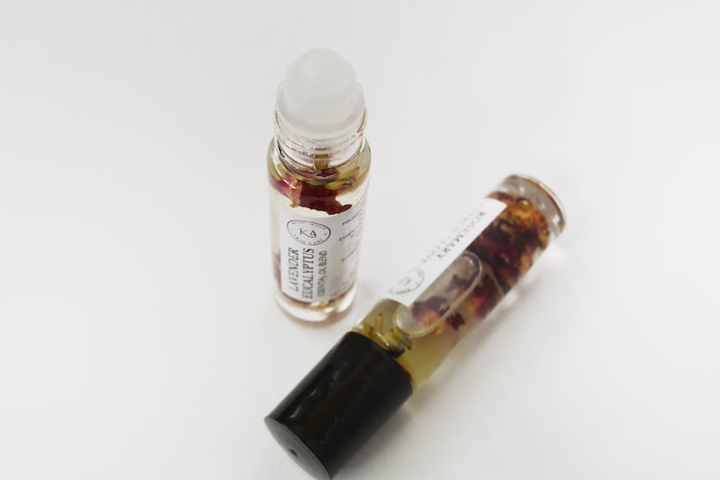 Pode incluir: Dois frascos roll-on de &oacute;leo essencial de vidro transparente com tampas pretas e r&oacute;tulos brancos. Um frasco est&aacute; em p&eacute;, o outro est&aacute; inclinado. Os frascos cont&ecirc;m um l&iacute;quido transparente com p&eacute;talas de flores secas. O r&oacute;tulo do frasco vertical diz "Lavender Eucalyptus Essential Oil Blend."