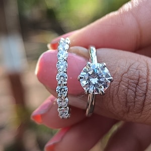 Puede incluir: Primer plano de un anillo de compromiso de plata con un gran diamante redondo y transparente. Una banda de plata con diamantes más pequeños se lleva en el mismo dedo.