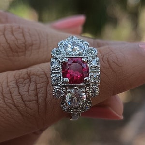 May include: A silver ring with a red gemstone surrounded by smaller clear gemstones.